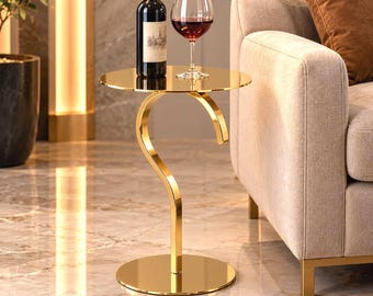 May include: A gold-coloured metal side table with a circular top and a curved, hook-shaped support stand on a round base. A dark glass wine bottle and a clear glass filled with red wine are placed on the reflective surface of the table.