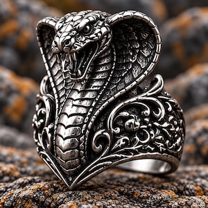 May include: A silver-toned ring featuring a detailed cobra head with an open mouth and flared hood. The band is decorated with intricate scrollwork and filigree patterns that extend from the base of the snake's neck along the sides of the ring.
