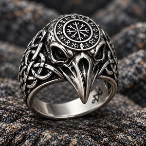 May include: A silver-toned ring featuring a detailed raven head design. The top of the head displays a circular emblem with a compass-like symbol surrounded by runic characters. The sides of the band are decorated with intricate, interwoven knotwork.
