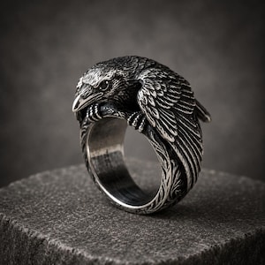 May include: A silver-toned metal ring featuring a detailed, three-dimensional raven perched atop the band. The bird has textured feathers and a sharp beak, while the band itself displays intricate, carved patterns along its outer surface.