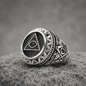 Op de afbeelding: Een zilveren ring met een cirkelvormig voorvlak met een verhoogde driehoek die een donkerrode stenen oog bevat. De band toont ingewikkelde gravures van een winkelhaak en passer aan de zijkant, omringd door gedetailleerde bladpatronen.