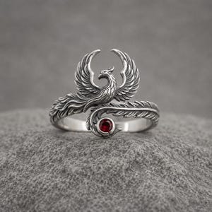 May include: A silver ring featuring a detailed phoenix design with wings spread upward. The bird's tail feathers wrap around the band, culminating in a small, round, deep red gemstone set at the end of the tail, creating a continuous metal loop.