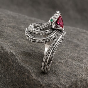 Könnte beinhalten: Ein silberner Ring in Form einer gewundenen Schlange mit texturierter, perlenartiger Oberfläche. Der Schlangenkopf ist mit einem dreieckigen roten Edelstein und kleinen grünen Edelsteinaugen besetzt. Die Ringschiene bildet den Schlangenkörper.