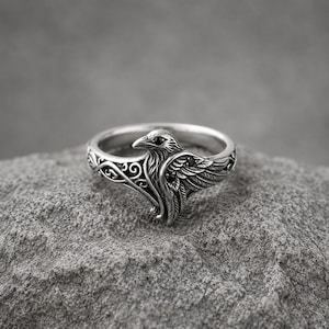 May include: A silver ring featuring a detailed raven design with engraved feather textures and scrollwork patterns. The bird's eye and wing are accented with small, dark, round stones, creating a contrast against the polished metal finish.