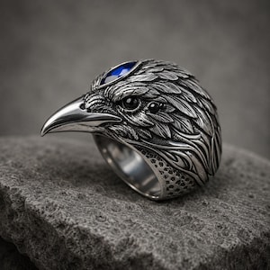 May include: A silver-toned metal ring shaped like a raven head with intricate feather detailing, a sharp beak, and a single marquise-cut blue gemstone set on the top of the head. The eye of the bird is a small, round, dark black stone.