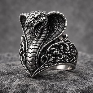 May include: A silver-toned ring featuring a detailed cobra head with an expanded hood and open mouth showing fangs. The band is decorated with intricate scrollwork and relief patterns, showcasing a metallic finish with dark oxidised accents.