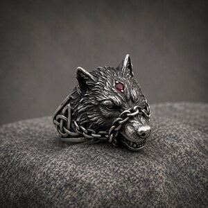 May include: A silver-coloured ring featuring a detailed wolf head sculpture with a small red gemstone set on the forehead. The wolf is adorned with a thin metal chain wrapped around its snout, and the band displays intricate knotwork patterns.
