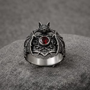 Puede incluir: Un anillo de tono plateado con un motivo detallado de murciélago con las alas extendidas. El centro del anillo muestra una piedra preciosa redonda de color rojo intenso facetado, flanqueada por dos piedras negras más pequeñas sobre un fondo de filigrana de metal oscuro.