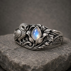 May include: A silver ring with an owl and a teardrop-shaped gemstone. The ring has detailed floral and leaf patterns. The gemstone has a blue and white iridescent glow. The ring is displayed on a grey stone surface.