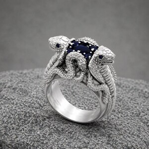 Può includere: Un anello in argento con due teste di serpente dettagliate rivolte verso l'esterno, i cui corpi si avvolgono attorno a un gruppo centrale di gemme blu scuro dal taglio ovale. La fascia in metallo ha una finitura lucida e squame finemente lavorate.