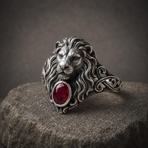 Könnte beinhalten: Ein silberfarbener Ring mit einer detaillierten Löwenkopf-Skulptur und fließender Mähne. Unter dem Kinn des Löwen ist ein ovaler, tiefroter facettierter Edelstein in das Metall eingefasst. Die Ringschiene zeigt komplexe, geschwungene Ornamente.