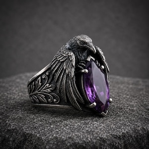 May include: A silver-toned metal ring featuring a detailed raven sculpture with spread wings. The bird holds a large, marquise-cut purple gemstone in its talons. The band of the ring is decorated with intricate, embossed leaf-like scrollwork patterns.