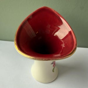 以下が含まれることがあります： 三角形の開口部を持つ陶器製の花瓶で、内側は深い赤色、縁は金色で縁取られています。外側はオフホワイトで、側面に花の模様が描かれています。花瓶は首が細く、上部に向かって広がっている形状をしています。