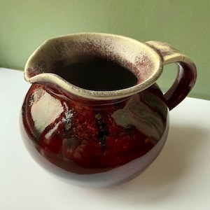 May include: A ceramic pitcher with a rounded body and a single handle. The exterior features a deep, glossy burgundy glaze with dark vertical streaks. The wide rim and interior are finished in a contrasting matte cream color with brown speckling.