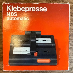 Puede incluir: Una máquina automática Klebepresse N8S negra y naranja vintage. La máquina tiene un cuerpo negro con un asa y un botón naranja. La caja es naranja con texto blanco que dice "Klebepresse N8S automatic".