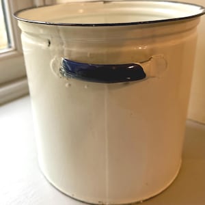 Może przedstawiać: Cylindryczne wiadro emaliowane w kolorze kremowym z ciemnoniebieskim uchwytem z boku oraz cienką ciemnoniebieską krawędzią u góry. Metalowy pojemnik ma gładkie wykończenie z widocznymi pionowymi szwami na całej powierzchni.