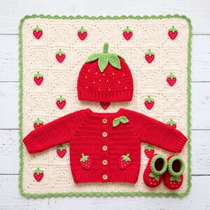 Può includere: Un cardigan rosso a maniche lunghe all'uncinetto con bottoni in legno e applicazioni a forma di fragola, un cappellino rosso abbinato a forma di fragola con foglie verdi e un paio di scarpine a fragola su una coperta all'uncinetto color crema.