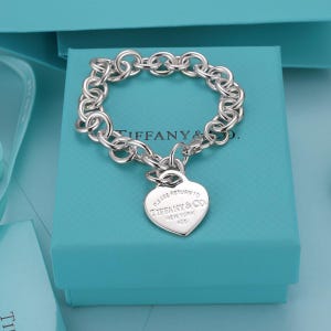 Puede incluir: Una pulsera de eslabones de plata de ley con un colgante en forma de corazón. El colgante tiene grabado el texto Please Return To Tiffany & Co. New York 925. La pulsera se presenta sobre un joyero cuadrado de color turquesa.