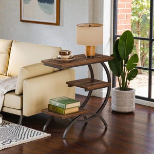May include: A three-tiered side table with a dark wood grain finish and a curved black metal frame. The top surface holds a beige lamp and a coffee cup on a saucer, while the bottom shelf supports two stacked books with green covers.