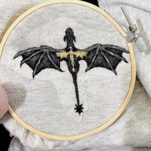 May include: Hand-stitched embroidery art of a dragon in a wooden hoop. The dragon features black wings and body with gold accents. The embroidery is on a light gray fabric. The wooden hoop is secured with a metal screw.