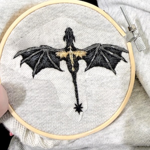 May include: Embroidered dragon design in a wooden hoop. The dragon is black with gold accents on its wings and chest, stitched on a gray fabric. The embroidery hoop is secured with a metal screw and clamp.