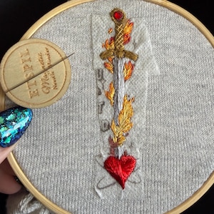 May include: An embroidery hoop containing light gray fabric with a stitched design of a silver sword engulfed in yellow and orange flames, topped with a red gem, piercing a red heart. A round wooden magnetic needle minder with the text ETSPIL is attached.