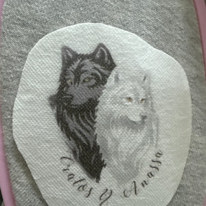 May include: A grey fabric patch featuring a printed illustration of two wolves, one black and one white, positioned side by side. Below the wolves, the text "Cratos y Anassa" is printed in a cursive script font on a white oval background.