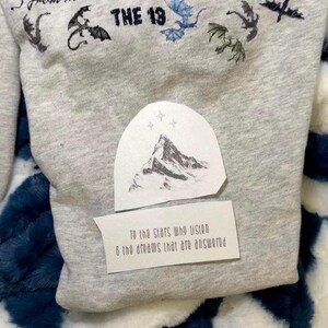 May include: A light gray heathered fabric featuring black and blue dragon illustrations, the text THE 13, a mountain graphic, and a white rectangular label printed with the text: To the stars who listen & the dreams that are answered.