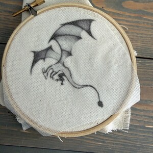 May include: A circular wooden embroidery hoop containing a piece of off-white fabric. Centered on the fabric is a black ink illustration of a winged dragon in flight, featuring a long tail with a pointed tip and detailed shading.