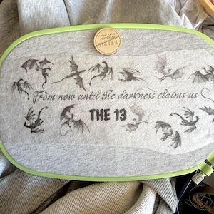 May include: A gray fabric stretched within a green embroidery hoop, featuring a dragon design and the text "From now until the darkness claims us THE 13." A small wooden Etsy logo is pinned to the fabric.