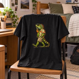 May include: A black short-sleeved t-shirt featuring a graphic print of a green iguana wearing black sunglasses, a floral print button-down shirt, and khaki shorts. The iguana is depicted in a walking pose with its tail extended behind it.