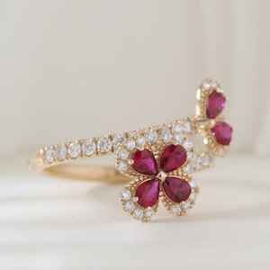 Pode incluir: Anel de ouro com design floral. Apresenta quatro pétalas de rubi rodeadas por um halo de pequenos diamantes. A banda também é adornada com pequenos diamantes. Fundo branco suave.
