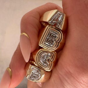 Pode incluir: Anéis dourados que soletram as letras A, B, C e D. Cada anel apresenta uma grande pedra preciosa transparente cortada em forma de letra. Os anéis são grossos e têm um acabamento polido.