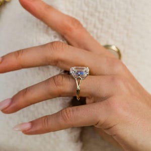 Puede incluir: Un anillo de oro con una gran piedra preciosa rectangular transparente y facetada en un engaste de cuatro garras. La banda presenta un diseño distintivo de hombro dividido que se estrecha en un solo hilo de oro fino en la base.
