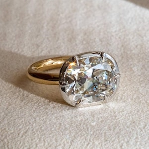 Puede incluir: Un anillo con una gran piedra preciosa transparente de talla cojín y facetas, montada en una estructura de garras de tono plateado. El aro del anillo está hecho de metal liso y pulido de tono dorado, creando un contraste entre ambos tonos.