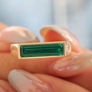 May include: A gold ring featuring a long, rectangular, baguette-cut green gemstone set horizontally in the center. The band has a smooth, polished finish, and a small, clear, round-cut stone is visible on the side of the setting.