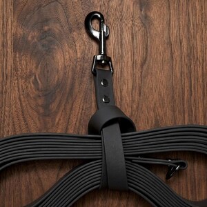 Op de afbeelding: Een zwarte hondenriem met een metalen sluiting en een leren lus. De riem is gemaakt van een plat, flexibel materiaal en is gelust en vastgebonden. De achtergrond is een donkere houtnerf.