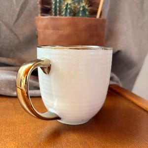 May include: A white ceramic mug with a gold-colored handle and rim. The mug has a ribbed texture and sits on a wooden surface. A brown leather pot with a cactus is in the background.