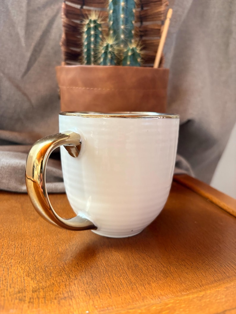May include: A white ceramic mug with a gold-colored handle and rim. The mug has a ribbed texture and sits on a wooden surface. A brown leather pot with a cactus is in the background.