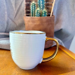 May include: White ceramic mug with a gold handle and rim. The mug has a ribbed texture and a gold inscription. A small cactus in a brown leather pot is in the background.