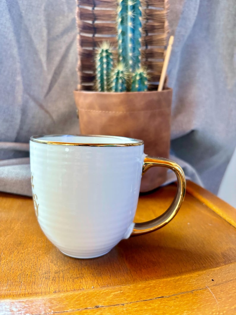 May include: White ceramic mug with a gold handle and rim. The mug has a ribbed texture and a gold inscription. A small cactus in a brown leather pot is in the background.