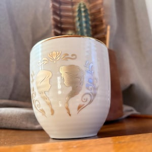 May include: Cream-colored ceramic cup with gold trim and gold silhouette designs of two women with long braids. The cup has a rounded shape and is decorated with floral and swirl accents. The background includes a cactus and a woven basket.