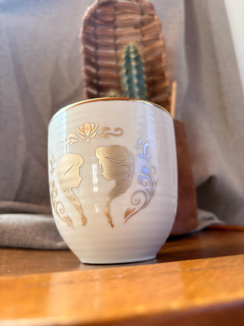 May include: Cream-colored ceramic cup with gold trim and gold silhouette designs of two women with long braids. The cup has a rounded shape and is decorated with floral and swirl accents. The background includes a cactus and a woven basket.
