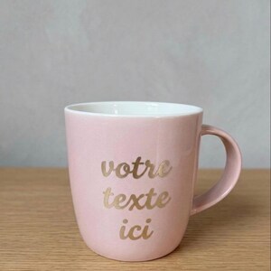 May include: A light pink ceramic mug with a white interior and a curved handle. The front of the mug features the gold-colored text "votre texte ici" written in a cursive script font, centered on the exterior surface of the vessel.