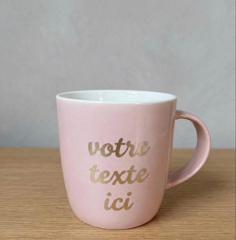 May include: A light pink ceramic mug with a white interior and a curved handle. The front of the mug features the gold-colored text "votre texte ici" written in a cursive script font, centered on the exterior surface of the vessel.