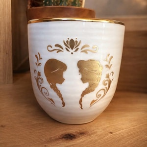 May include: White ceramic mug with a gold rim and gold silhouette designs. The mug features two female profiles and floral accents. The mug is likely used for drinking beverages and is a decorative item.
