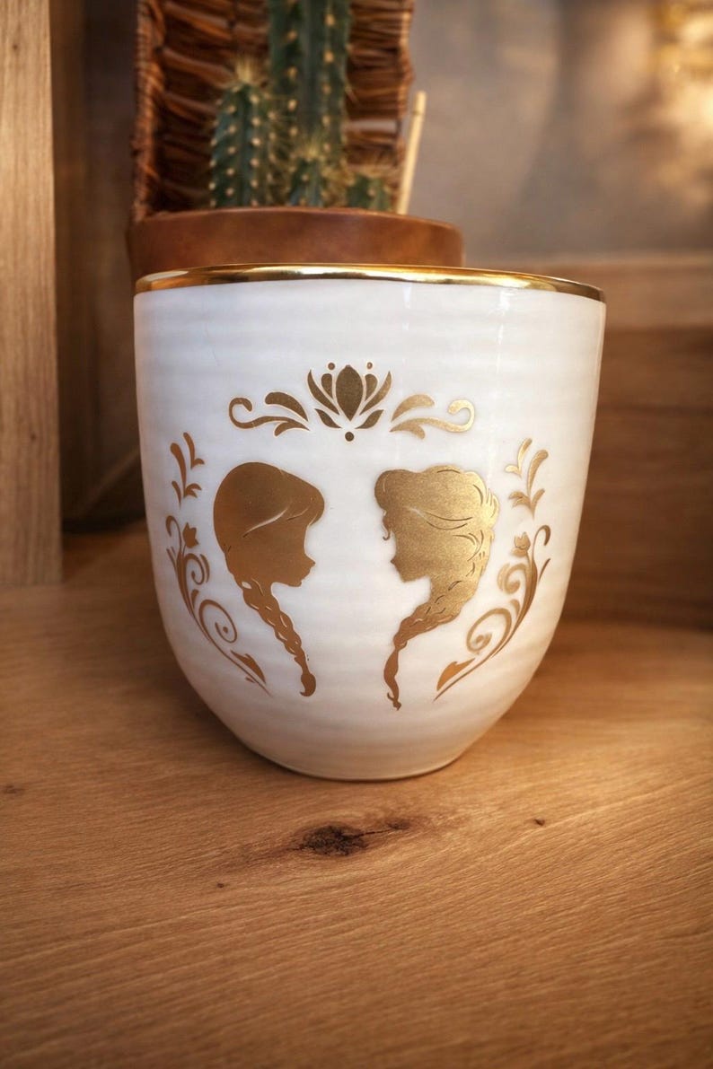 May include: White ceramic mug with a gold rim and gold silhouette designs. The mug features two female profiles and floral accents. The mug is likely used for drinking beverages and is a decorative item.