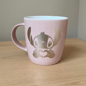 May include: A light pink ceramic mug featuring a metallic gold graphic of the character Stitch from Lilo and Stitch. The character is depicted with large ears and hands forming a heart shape. The interior of the mug is white and the handle is pink.