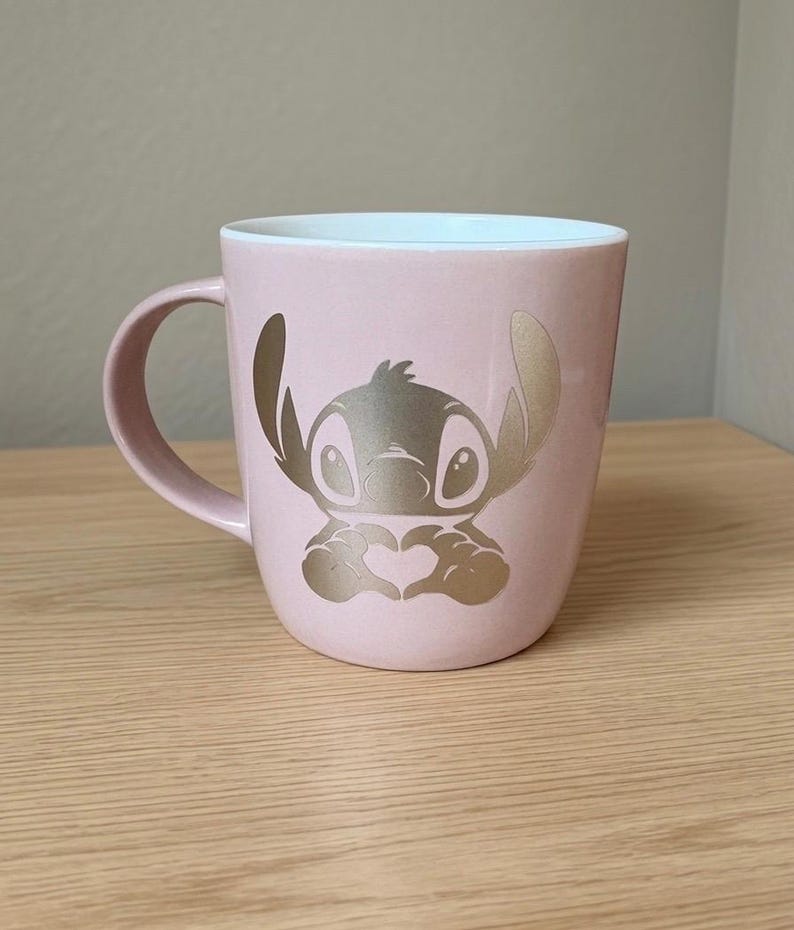 May include: A light pink ceramic mug featuring a metallic gold graphic of the character Stitch from Lilo and Stitch. The character is depicted with large ears and hands forming a heart shape. The interior of the mug is white and the handle is pink.