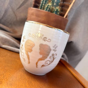 May include: A white ceramic mug with a gold rim and gold silhouette designs. The mug features two female profiles and decorative floral elements. The mug is set on a wooden surface.
